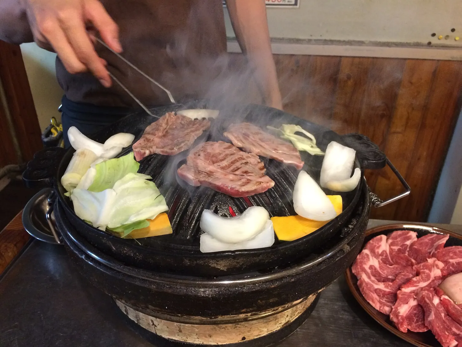Lamb and vegetables sizzling on a domed iron grill, turned with tongs, with a plate of raw lamb beside it