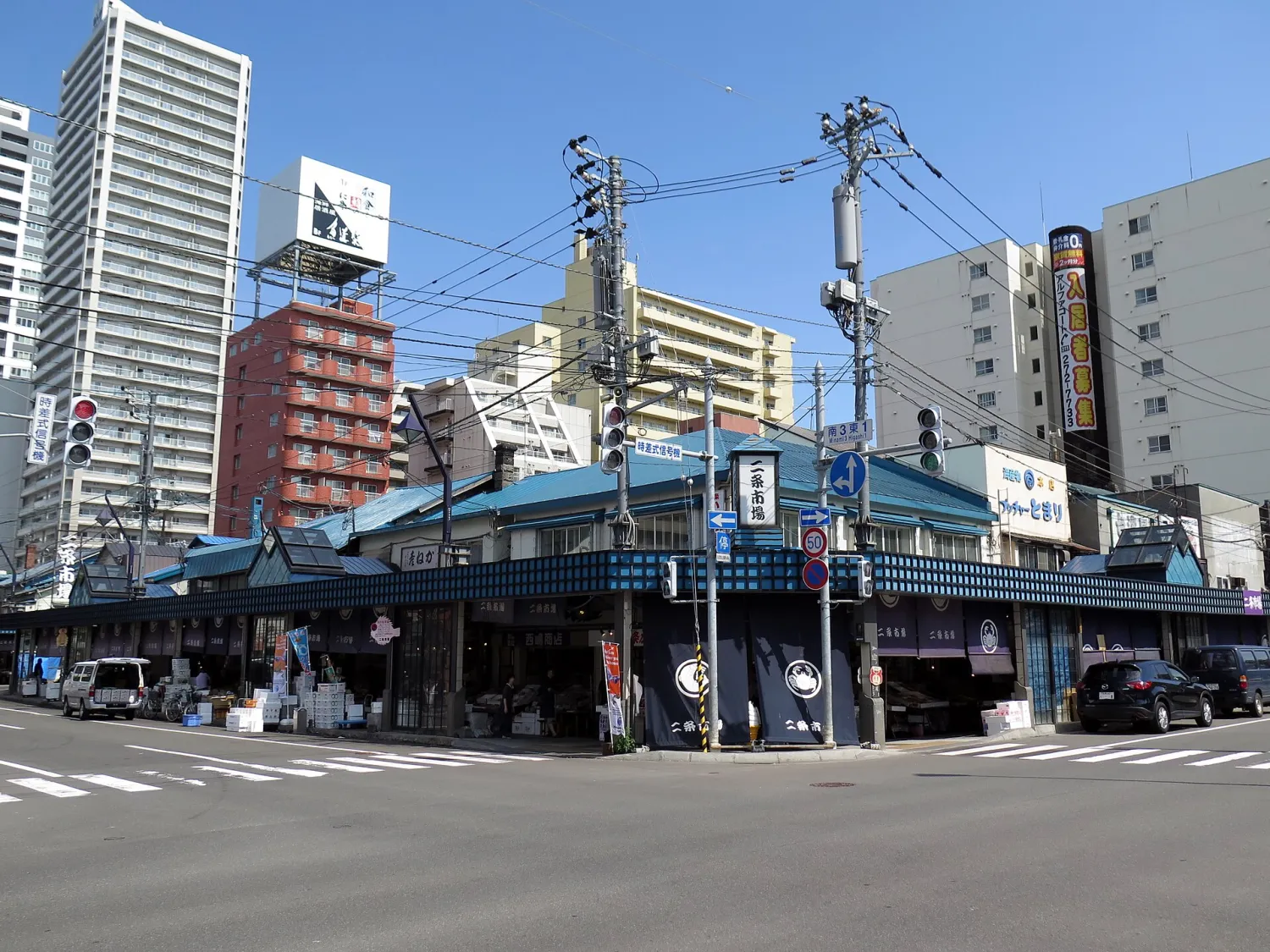 A low market building with blue awnings on a street corner, apartment and office blocks behind it