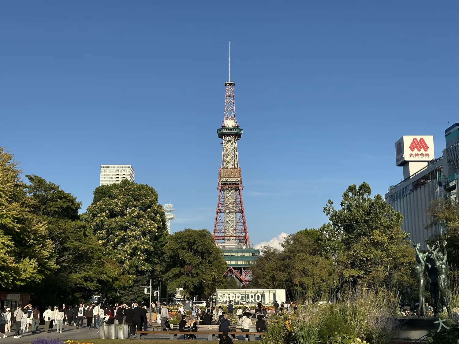 Ōdōri Park on a sunny day, trees and people on the paths, the Sapporo TV Tower at the far end