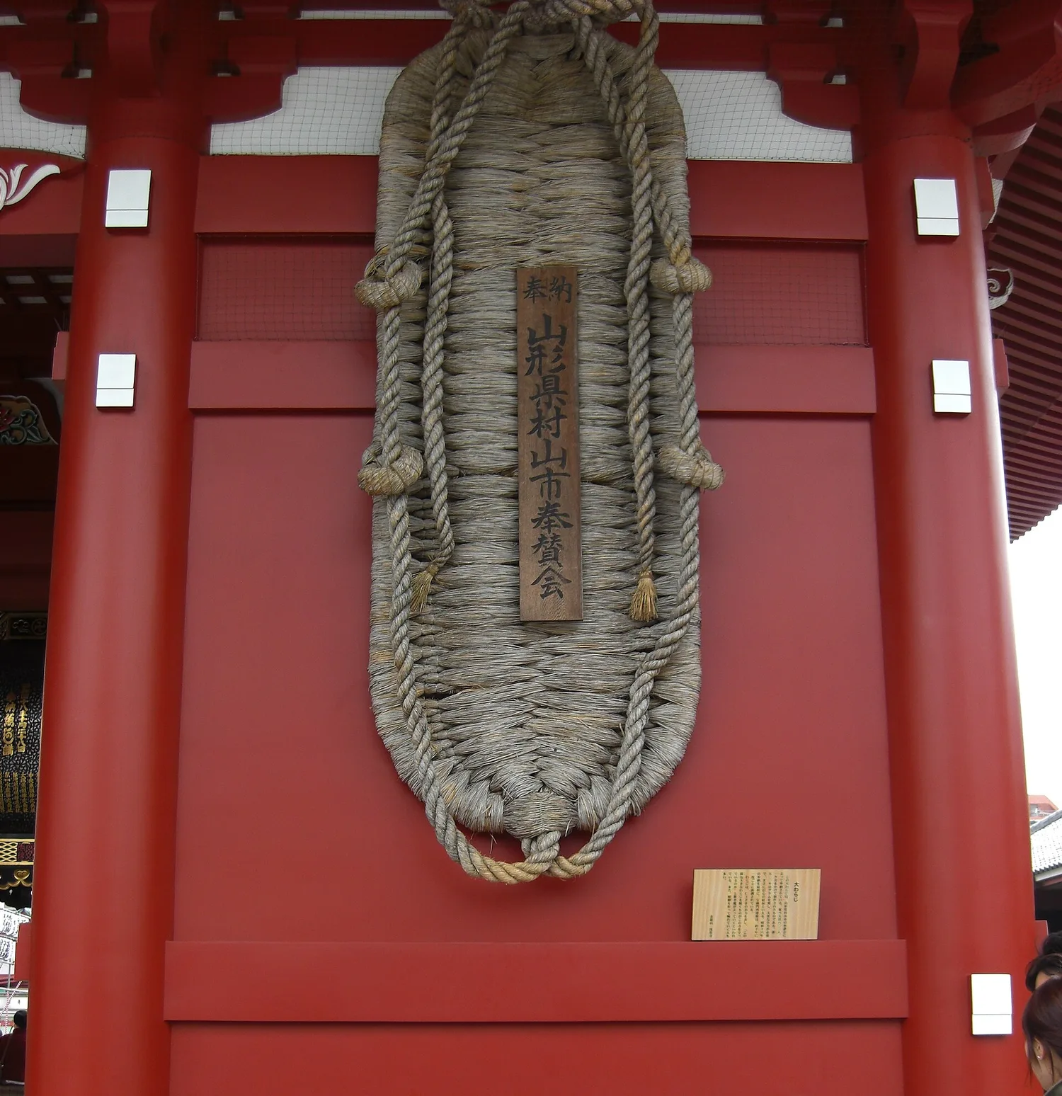 Giant straw sandal hanging on the red wall of Hozomon gate at Senso-ji, with a wooden plaque tied to it