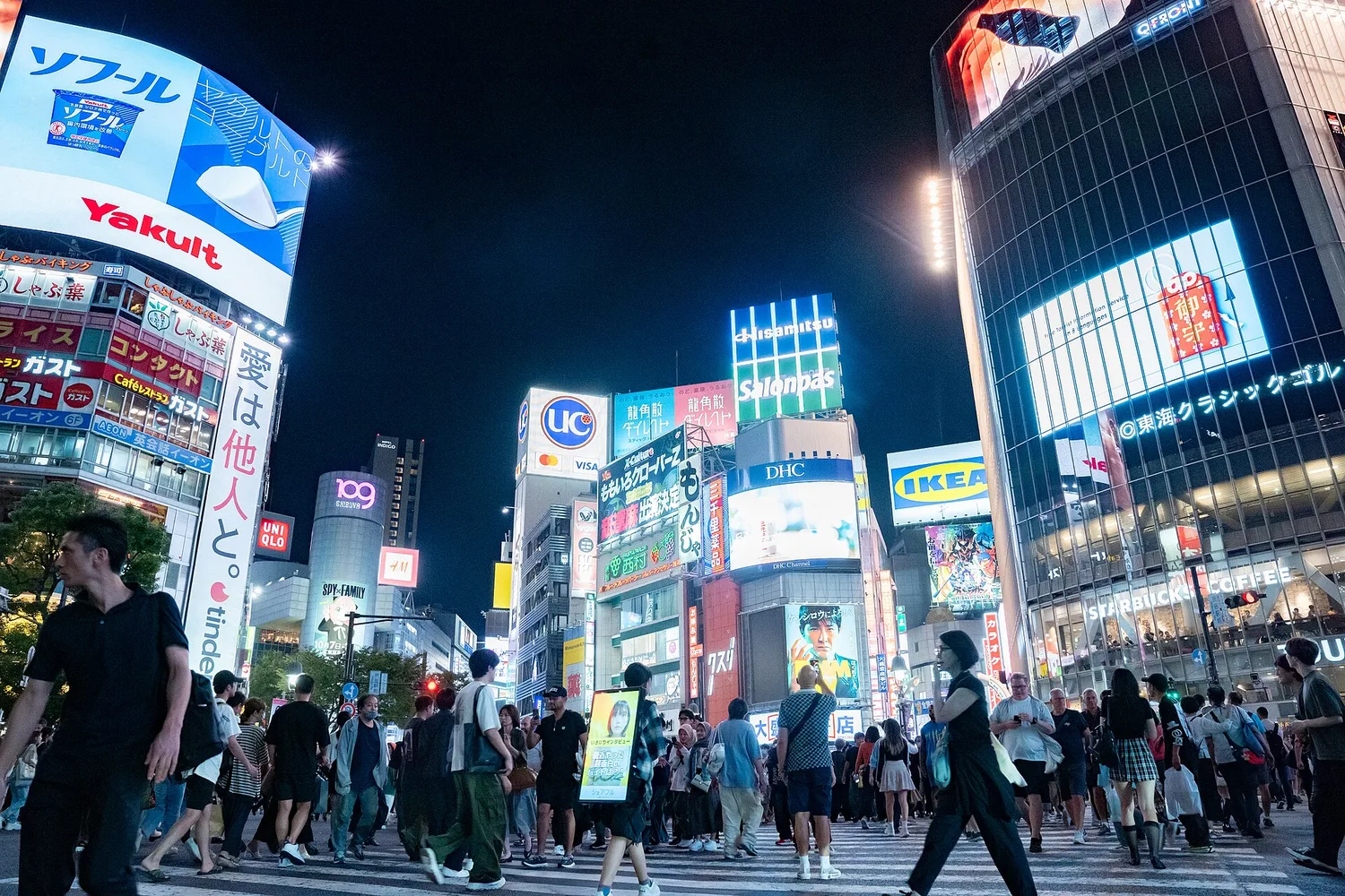 Shibuya Crossing at night from street level, with pedestrians crossing and bright signs on the buildings