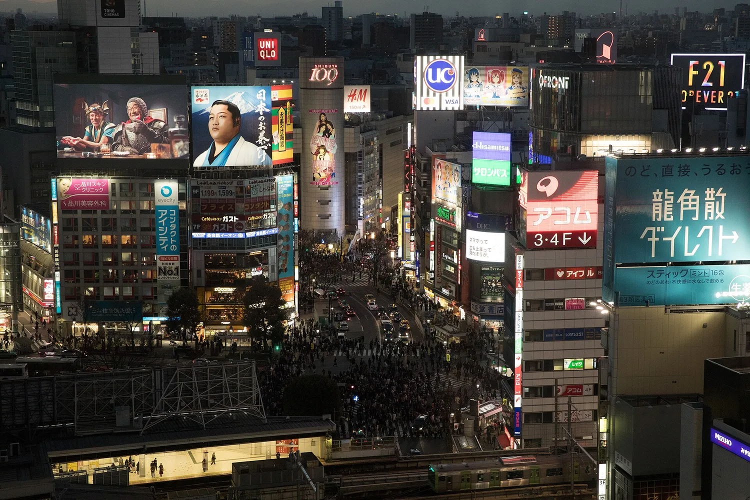 Shibuya Crossing at dusk seen from above, with large video screens on the buildings and crowds below