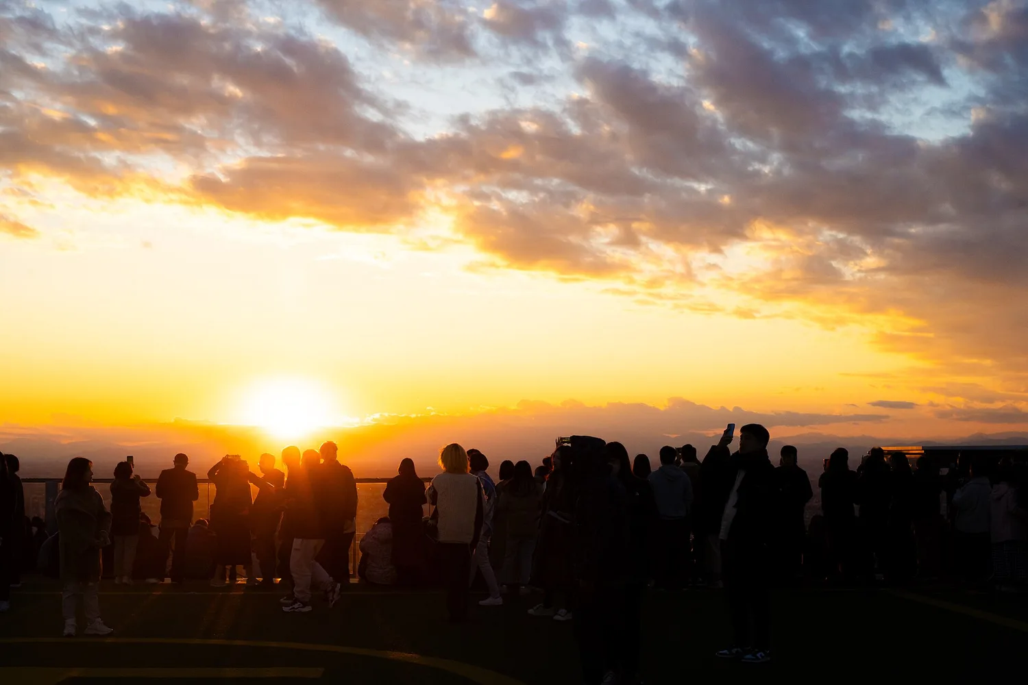 Visitors in silhouette facing the setting sun on the rooftop of Shibuya Sky, with orange clouds above