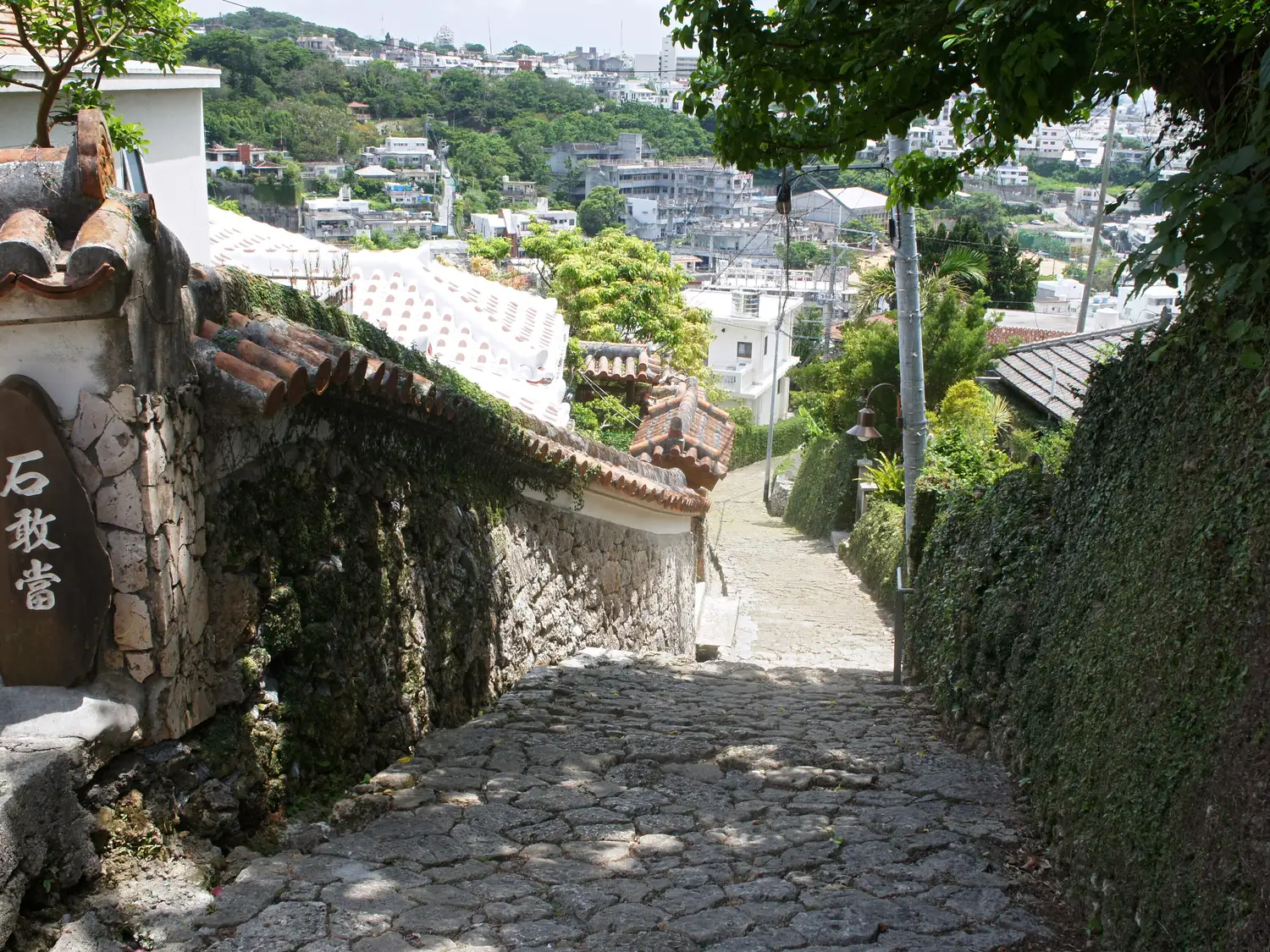 The Kinjo-cho stone path running downhill between a stone wall and a red-tiled house, the rooftops of Naha beyond