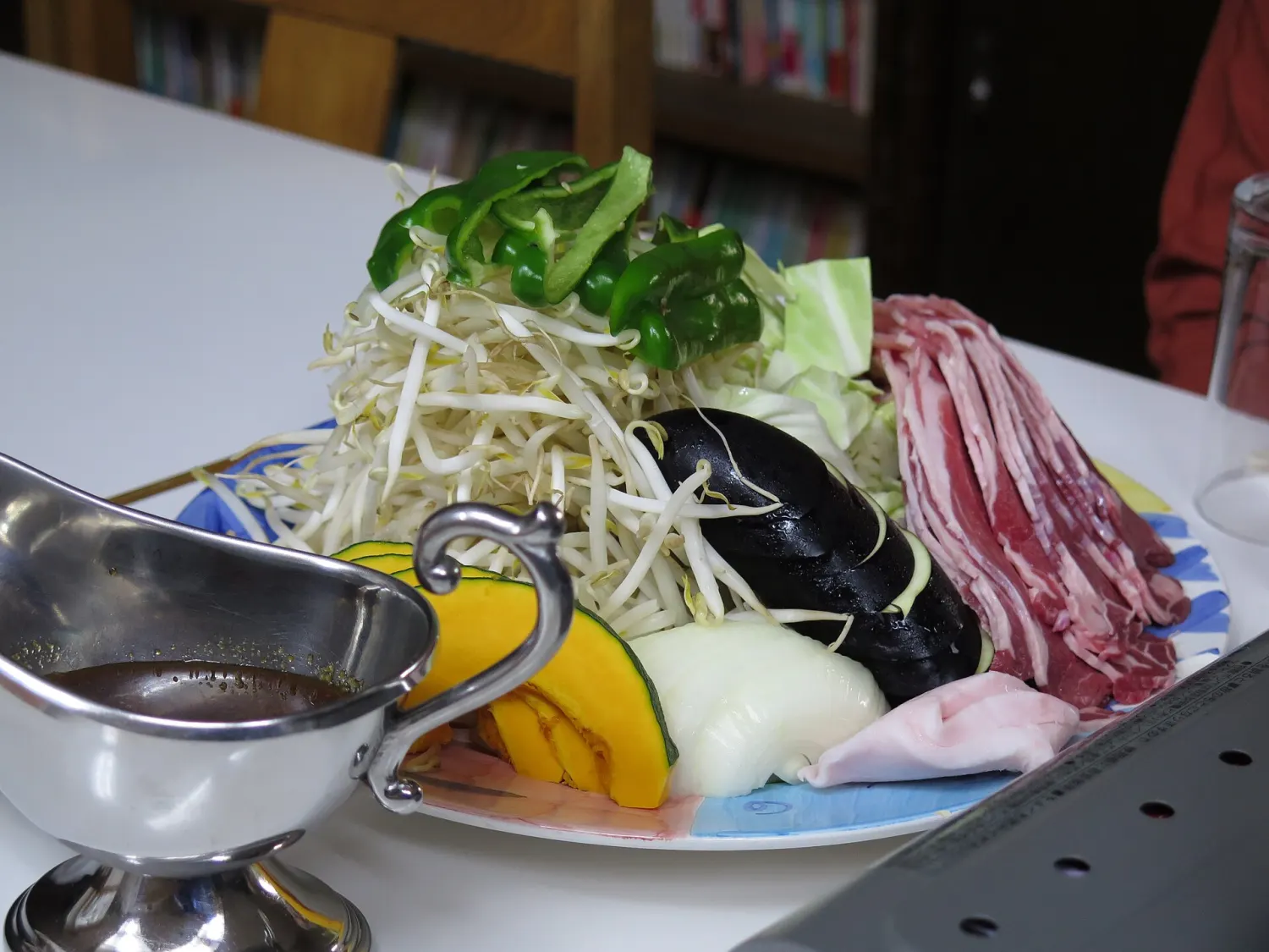 A plate piled with sliced lamb, bean sprouts, pumpkin, onion and green peppers beside a jug of dipping sauce
