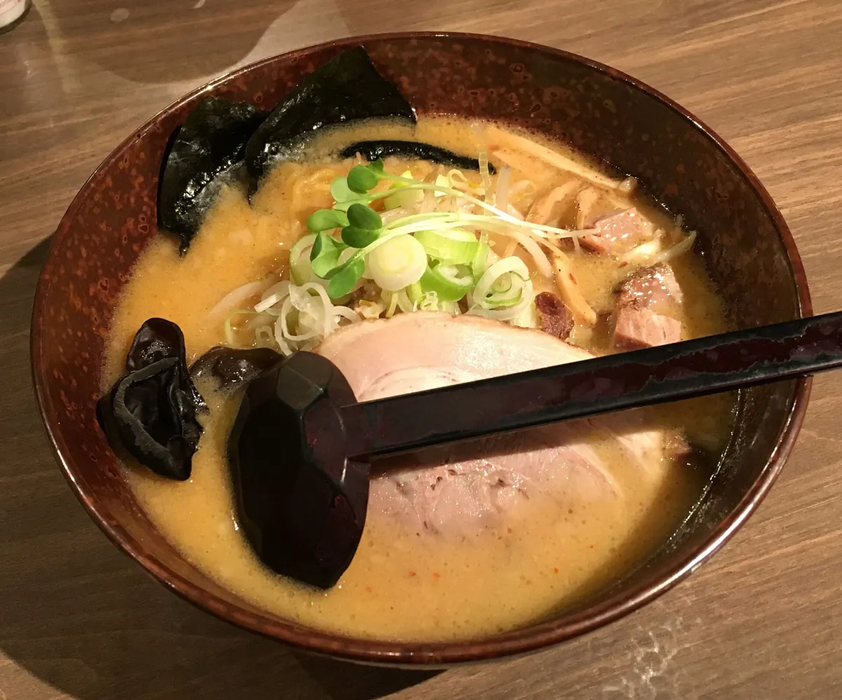 A dark bowl of miso ramen with a cloudy golden broth, sliced pork, spring onion, bean sprouts and wood-ear mushroom, a black ladle resting across it