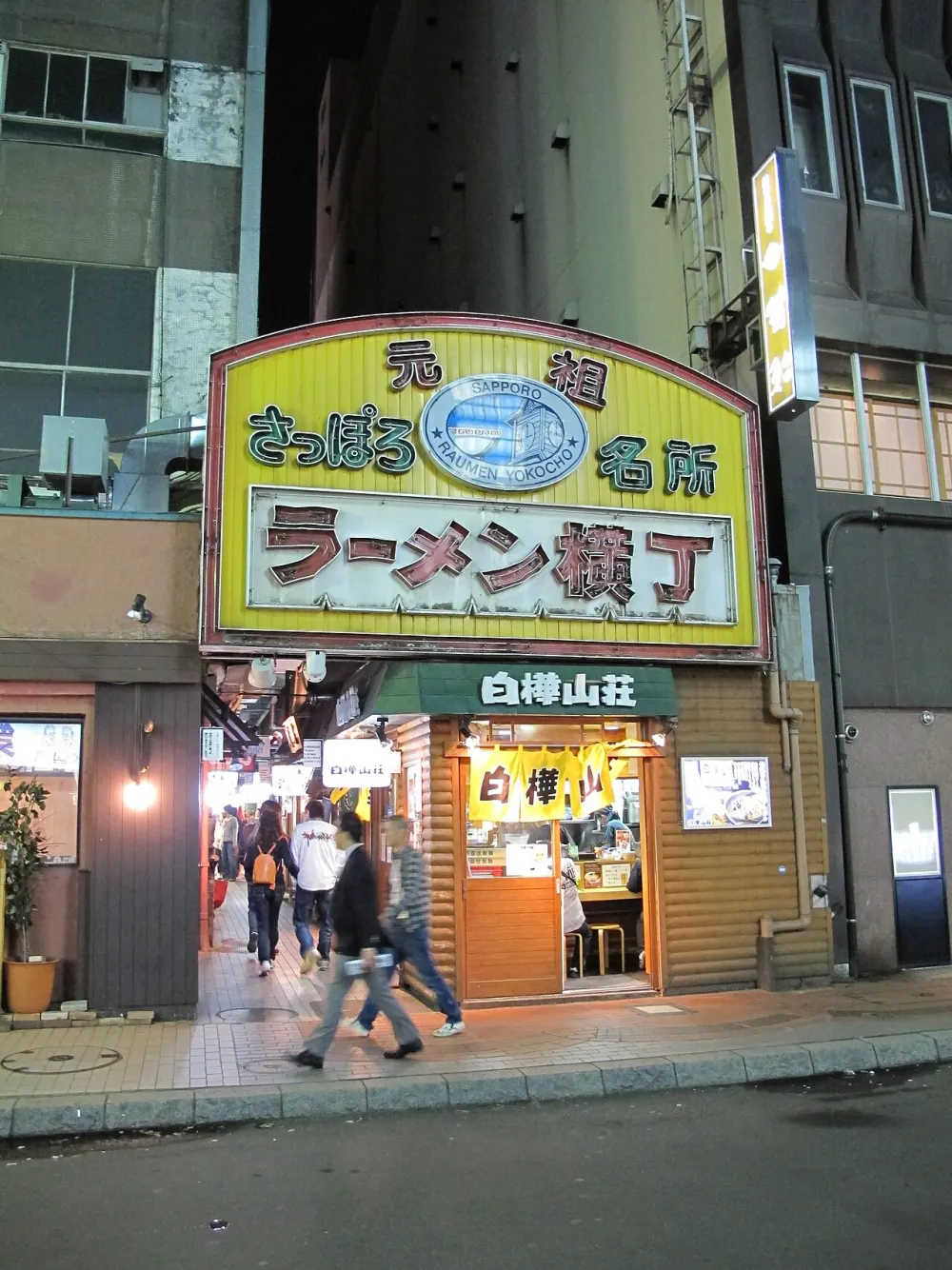 A lit yellow sign reading Ganso Sapporo Ramen Yokocho over a narrow entrance at night, people walking past