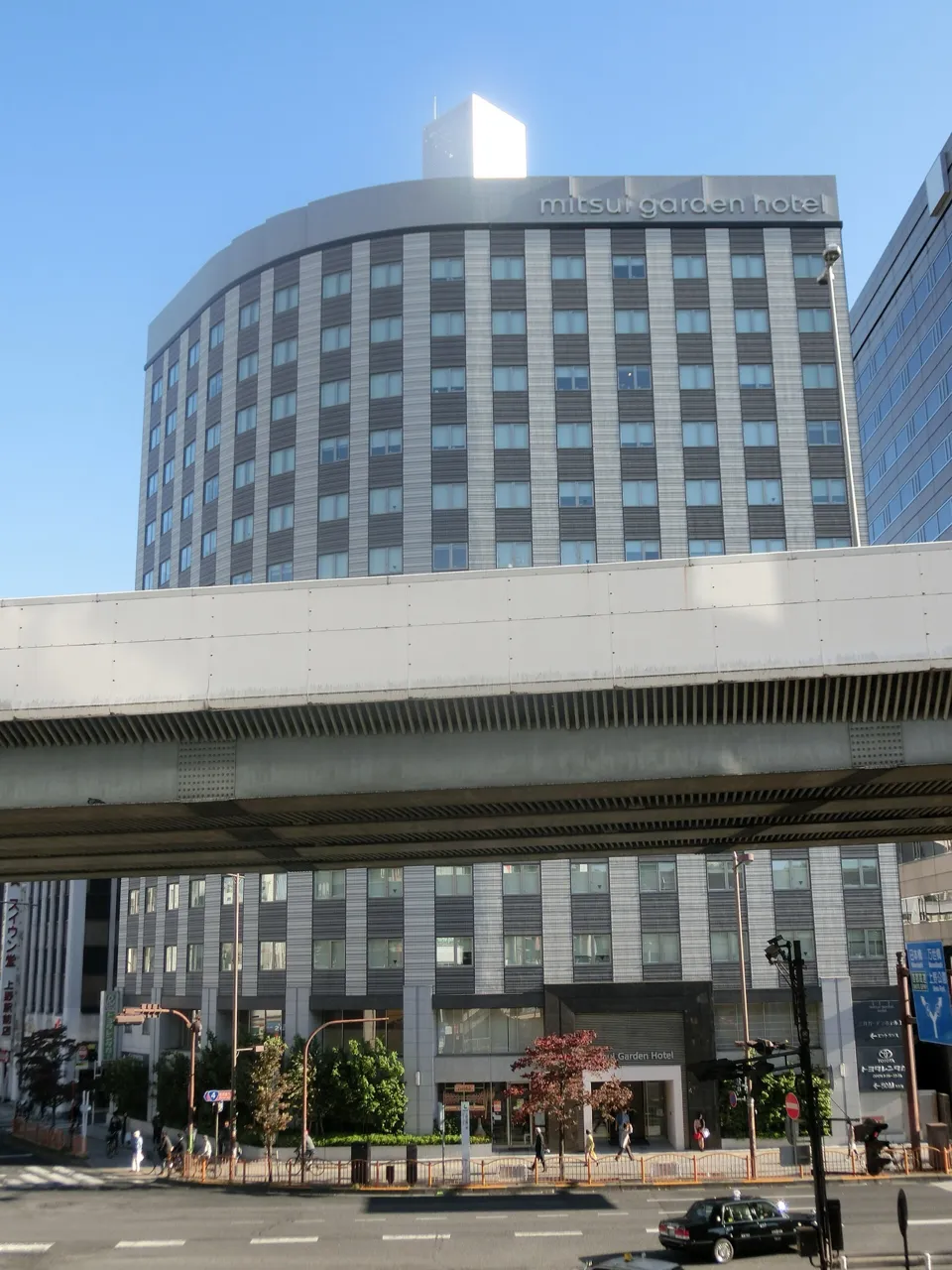 Mitsui Garden Hotel Ueno, a tall building with a curved roofline and the hotel's name at the top, behind an elevated road and a street corner with taxis