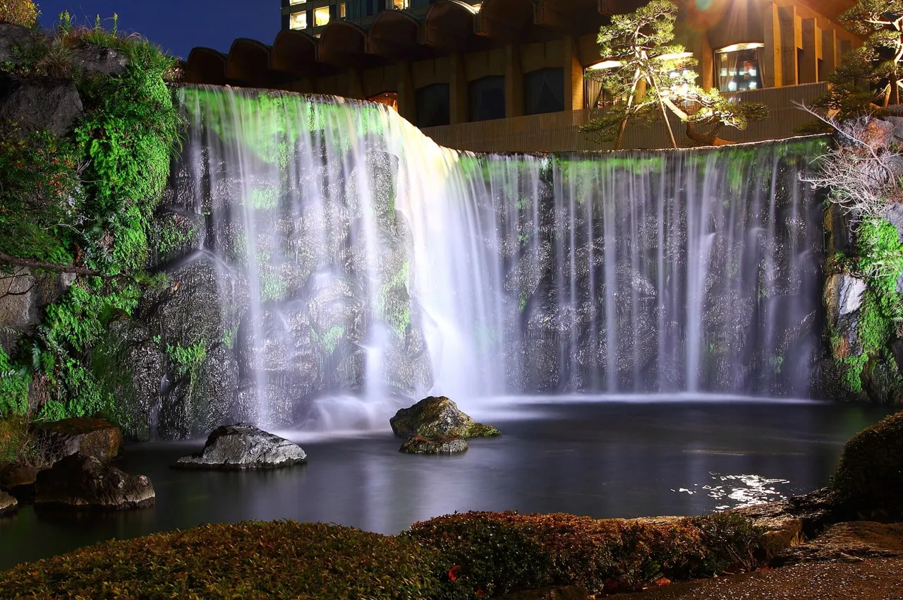 The waterfall in the Japanese garden of the Hotel New Otani Tokyo, lit up at night among dark rocks and trees