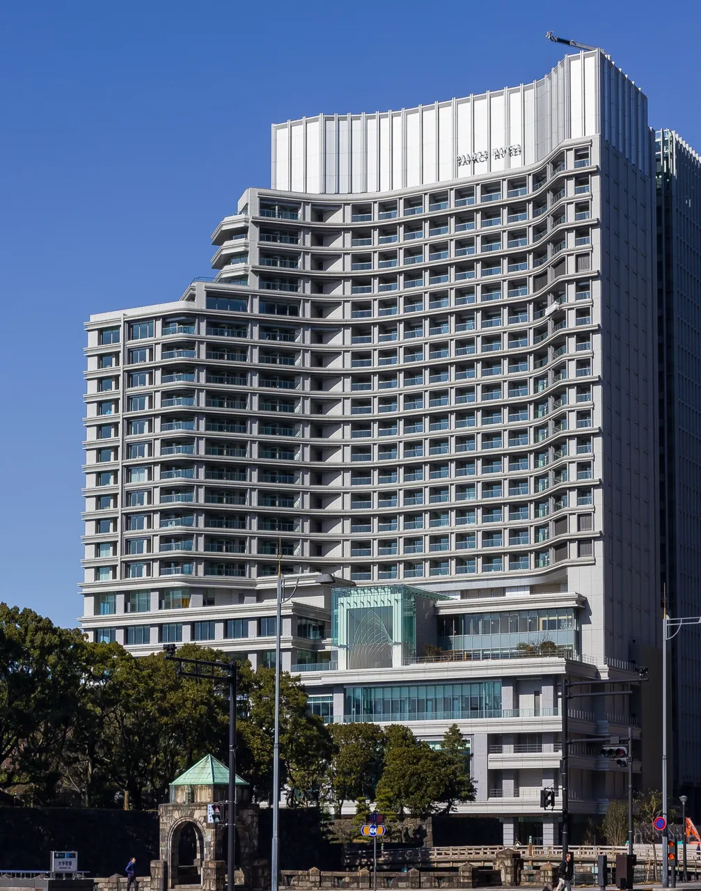 Palace Hotel Tokyo seen from the moat side of the Imperial Palace under a blue sky, with an old stone gate and greenery in the foreground