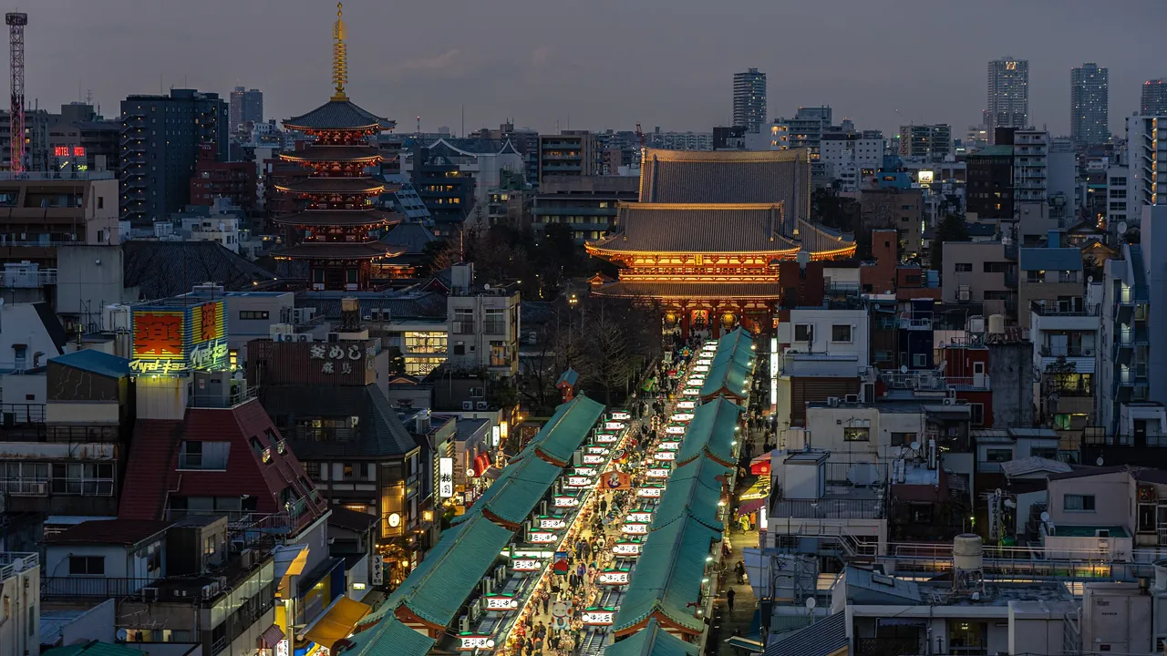 Senso-ji seen from above at dusk: the lit Hozomon gate and five-storey pagoda rise over the roofs of Asakusa, with the lit shopping street of Nakamise leading up to the gate