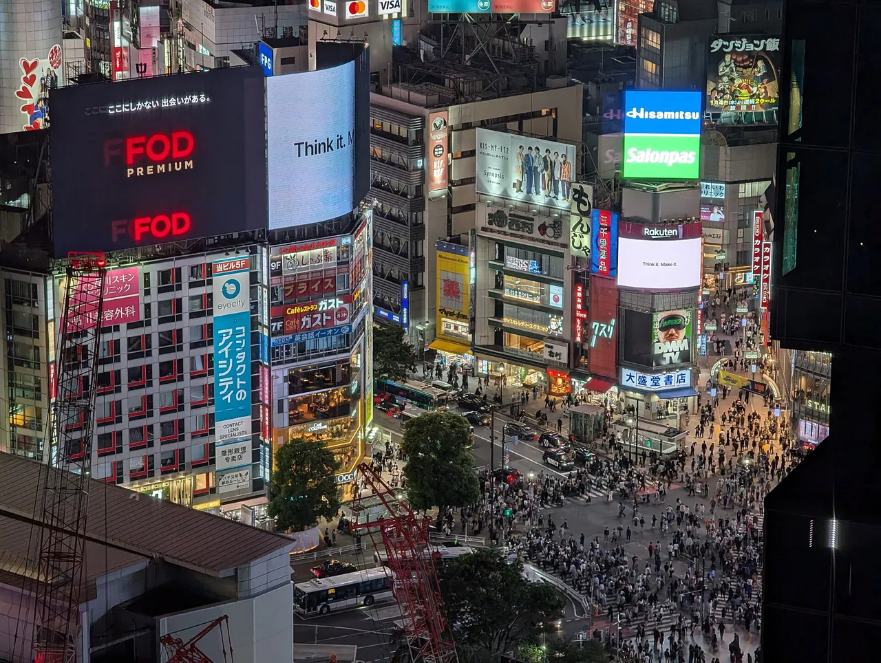 The Shibuya scramble crossing seen from high above in the evening, crowded with people, surrounded by buildings covered in lit screens and signs