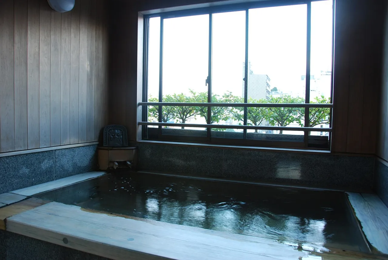 The communal bath of Ryokan Asakusa Shigetsu: a wooden tub and pale walls beside a wide window with green trees and buildings outside, in daylight
