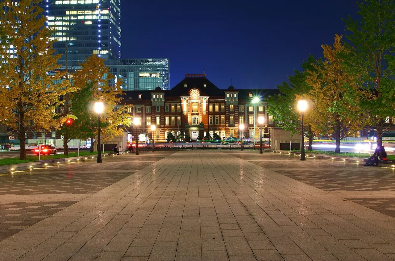 The red-brick Marunouchi building of Tokyo Station lit up at night, seen from the station plaza, with yellow autumn trees and street lamps on either side