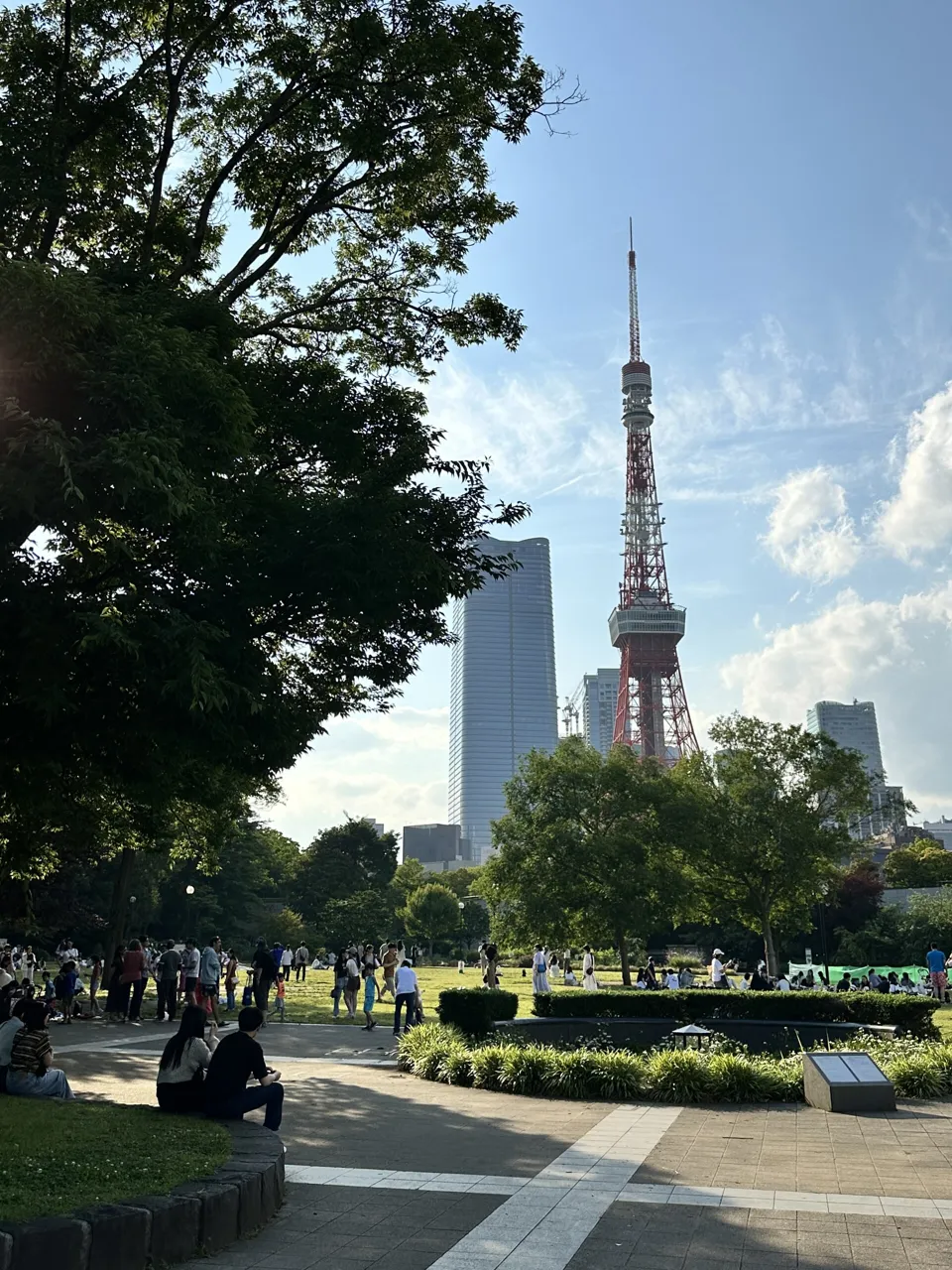 Tokyo Tower in red and white rising behind the tall green trees of Shiba Park on a sunny day, with people sitting on the lawn and paths below