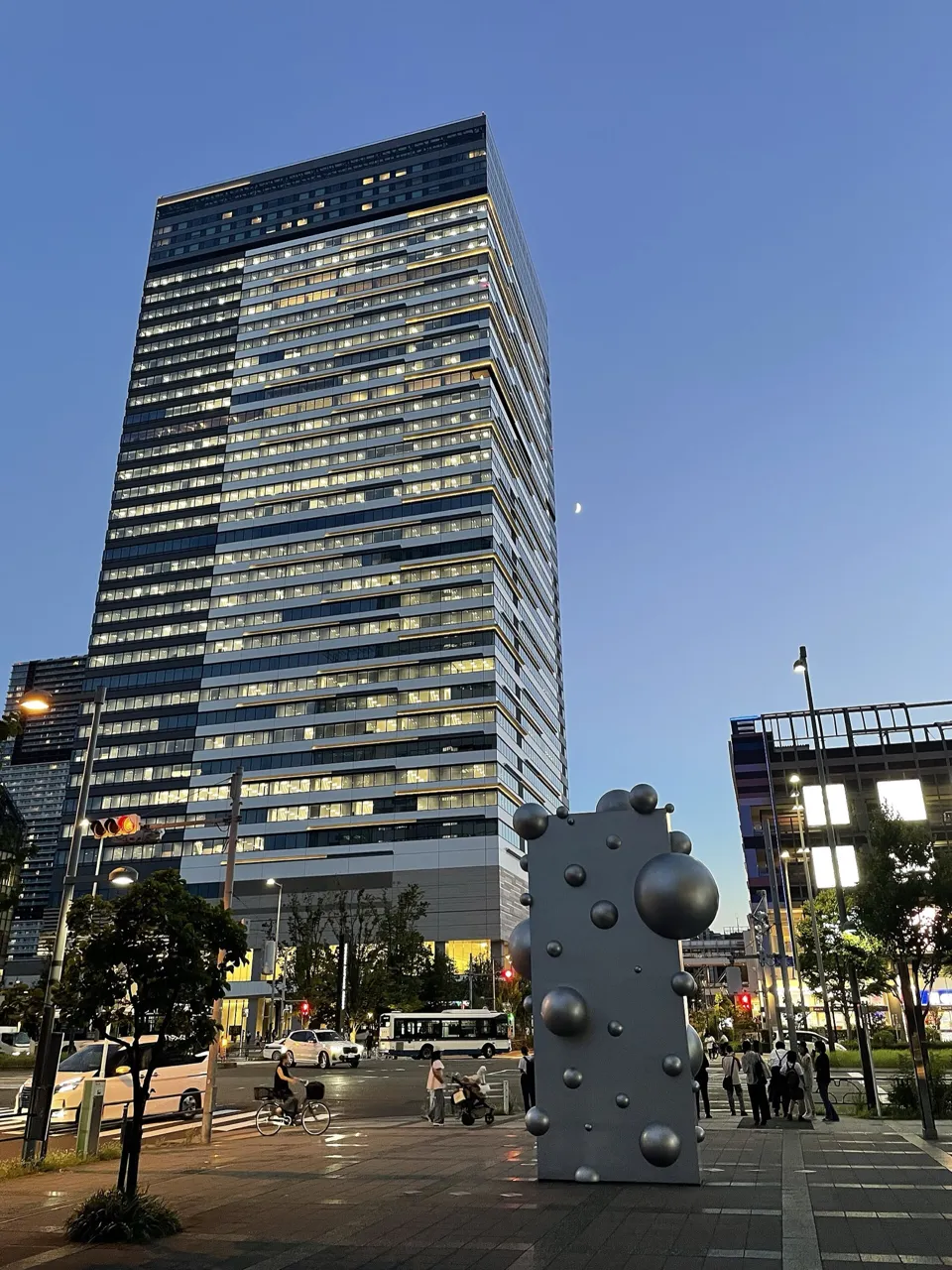 Toyosu Bayside Cross Tower at dusk, its windows lit and the moon in the sky, with a sculpture in the foreground; the hotel occupies the top floors