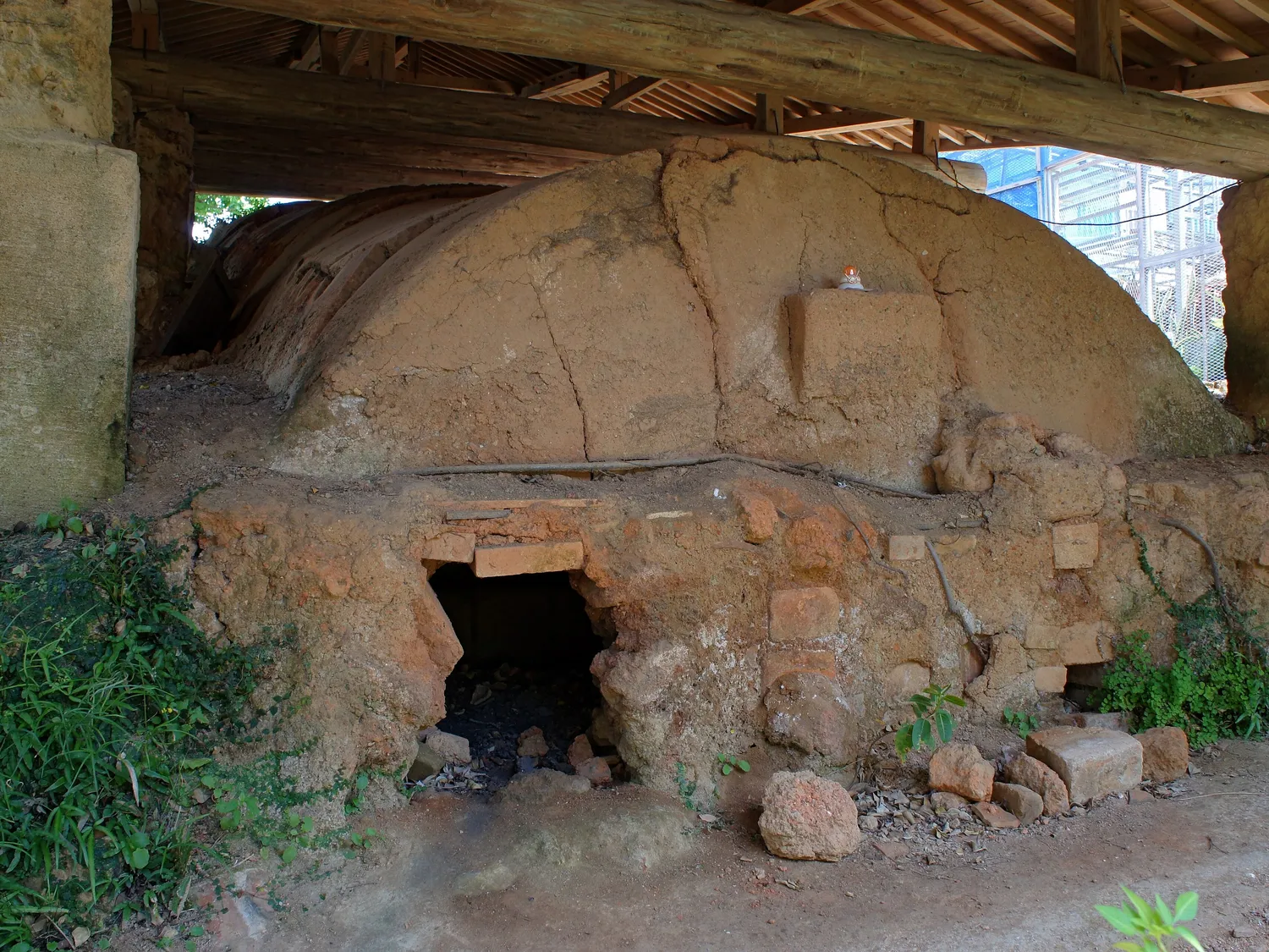 An old clay kiln with a rounded top and a small firing mouth, standing under a wooden roof