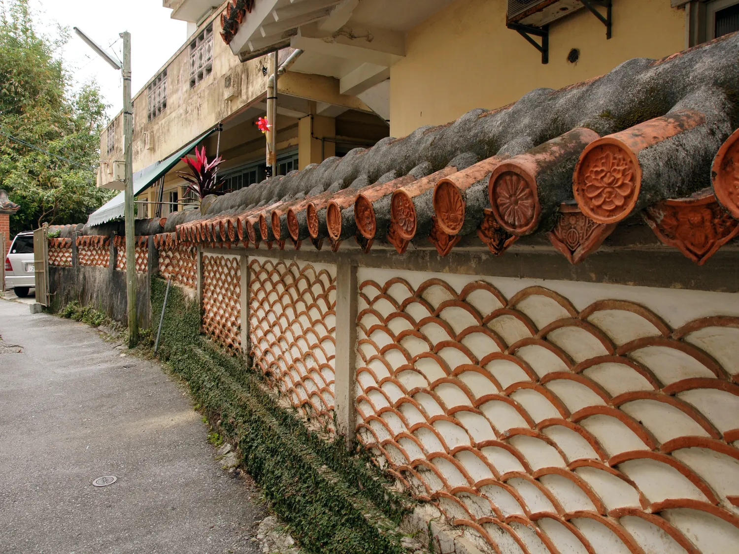 A lane-side wall faced with overlapping red roof tiles, with a row of rounded tile ends along the top