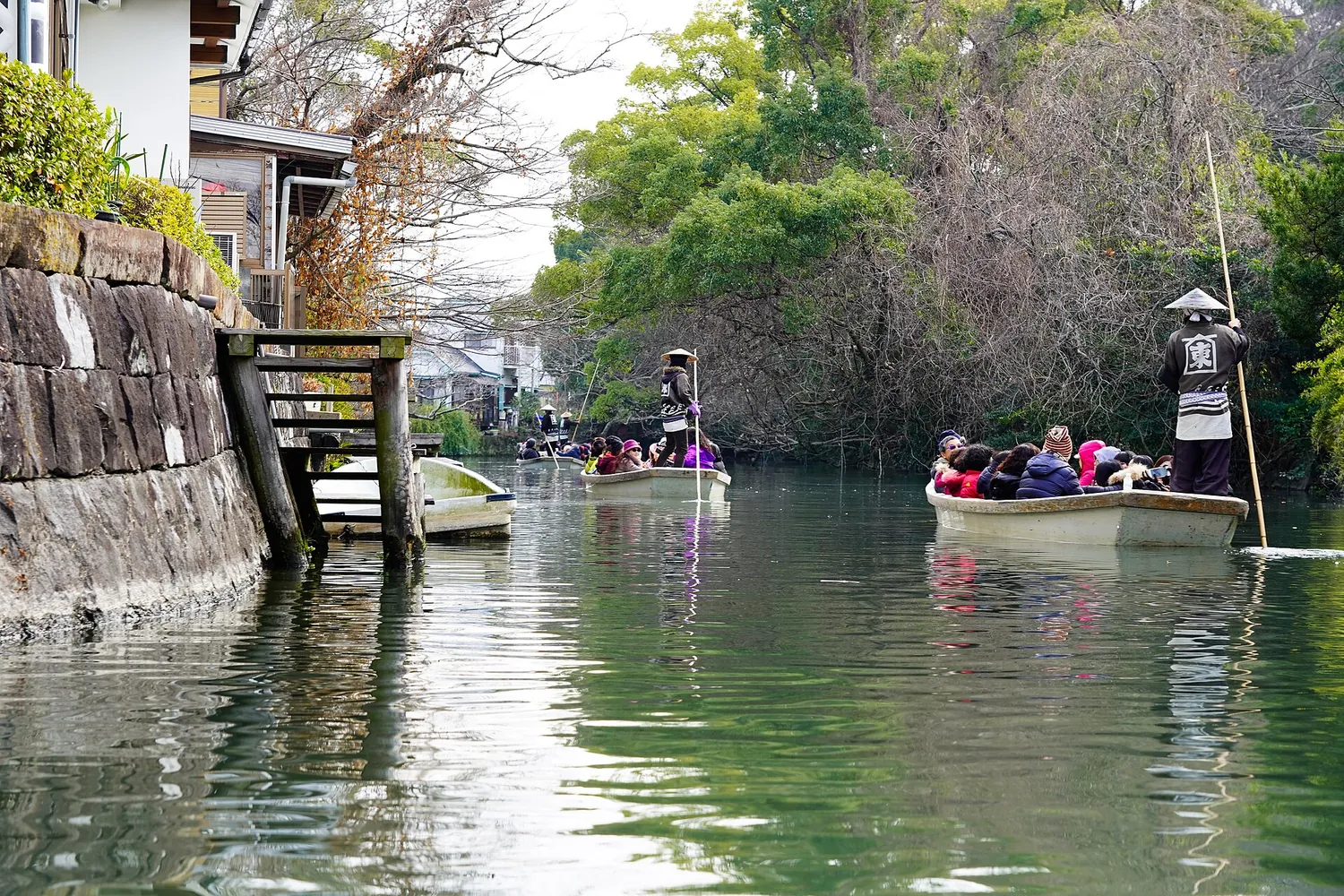 Passenger boats on a wide green canal, with a boatman standing with a long pole and a stone embankment alongside