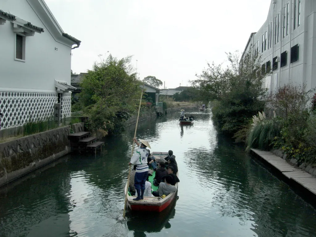 A boatman poling a small boat along a narrow canal beside a white building with a lattice-patterned lower wall