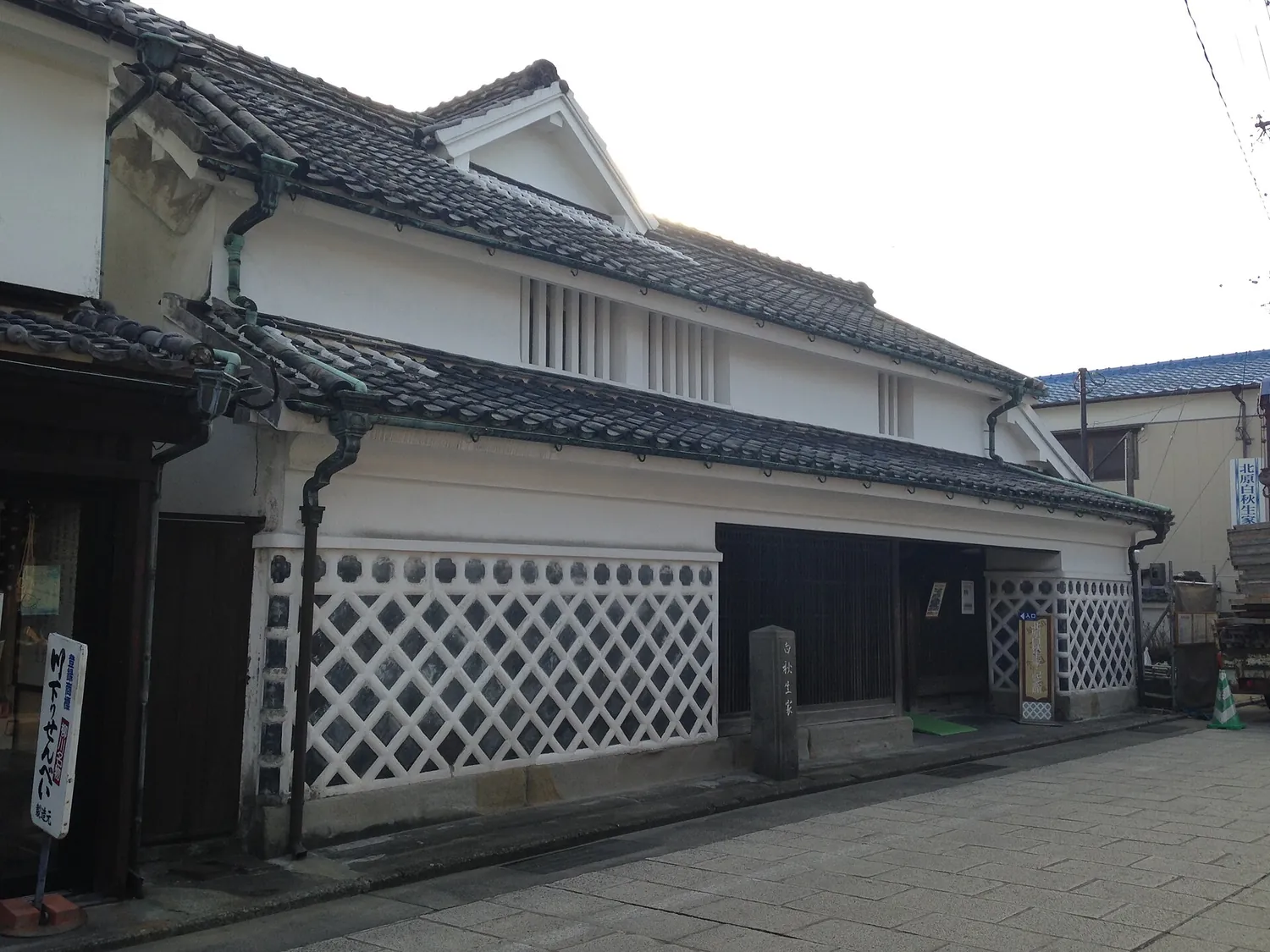 A white two-storey house with a dark tiled roof and a lattice-patterned lower wall on a quiet street